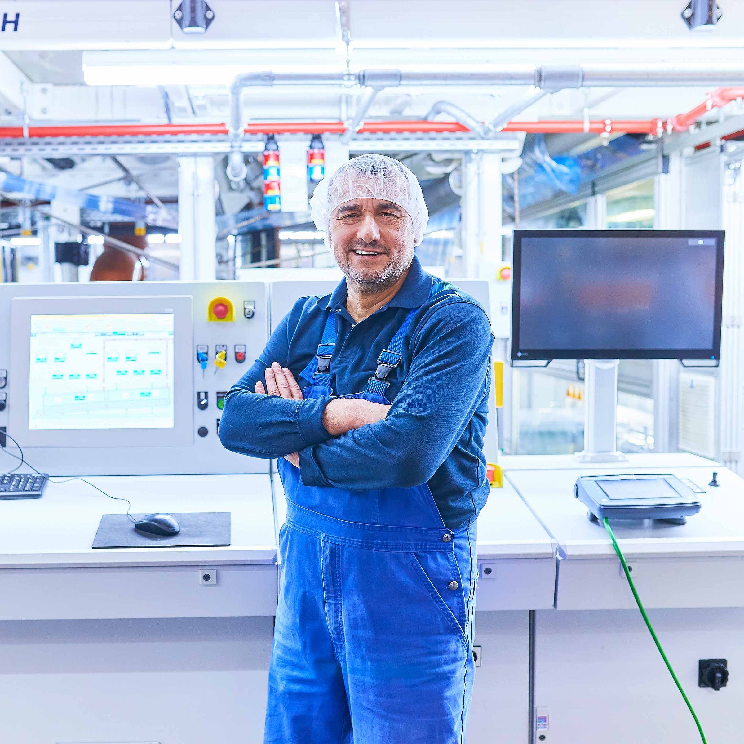 An employee wearing dark blue work clothes and a hairnet is standing in front of a machine on the production floor. He has his arms crossed and is smiling at the camera