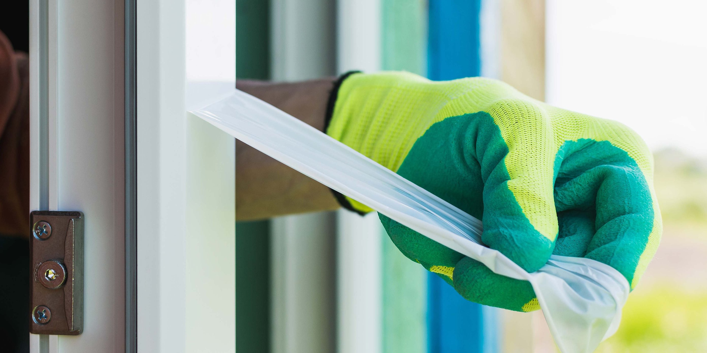 Eine Hand trägt einen grün-blauen Schutzhandschuh und zieht eine Oberflächenschutzfolie von einem Fensterrahmen ab