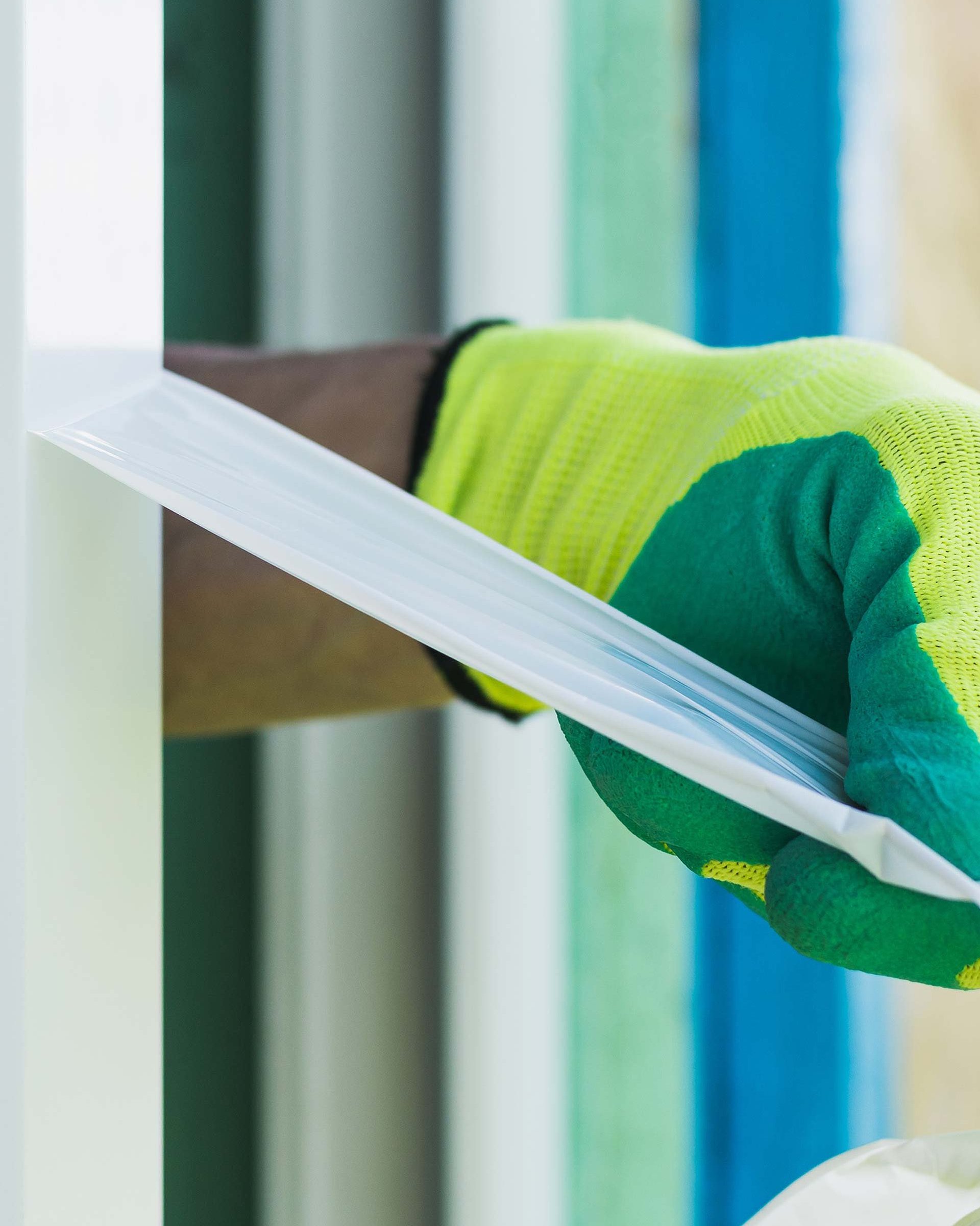 Eine Hand trägt einen grün-blauen Schutzhandschuh und zieht eine Oberflächenschutzfolie von einem Fensterrahmen ab
