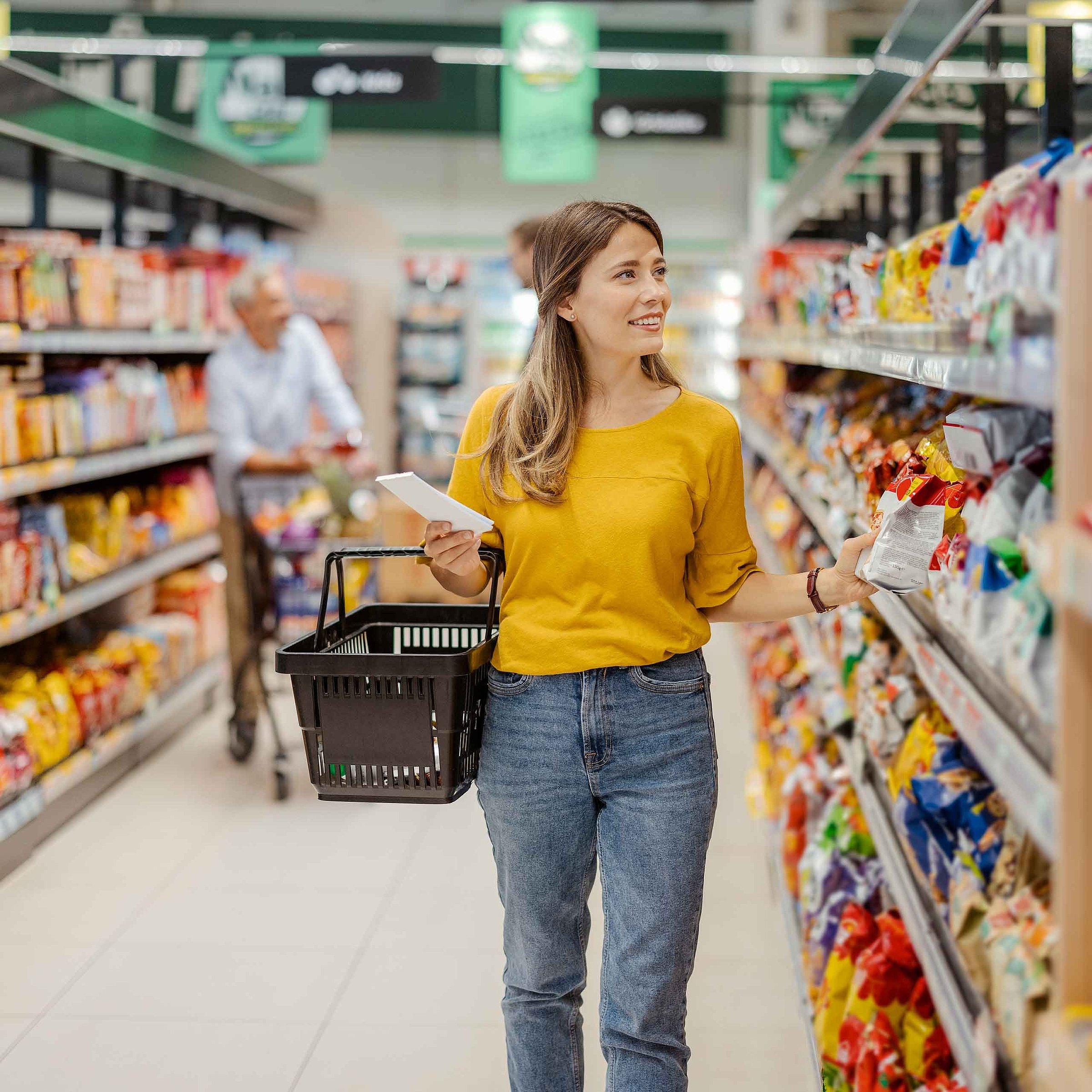A woman in a yellow T-shirt and jeans is in the supermarket holding a shopping basket. She is standing next to a shelf and has a shopping list in one hand and a bag of cookies in the other.