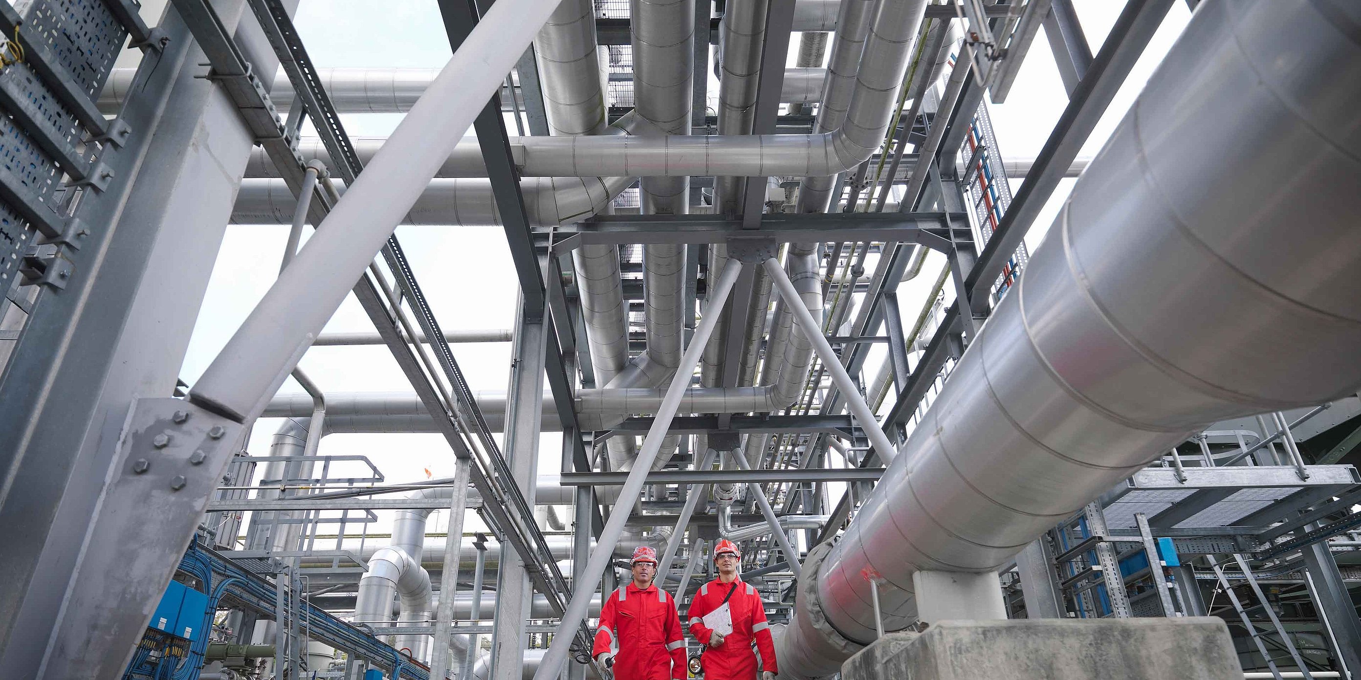Deux personnes portant des casques de protection et des vestes rouges dans un site industriel avec de nombreux tuyaux et des structures métalliques