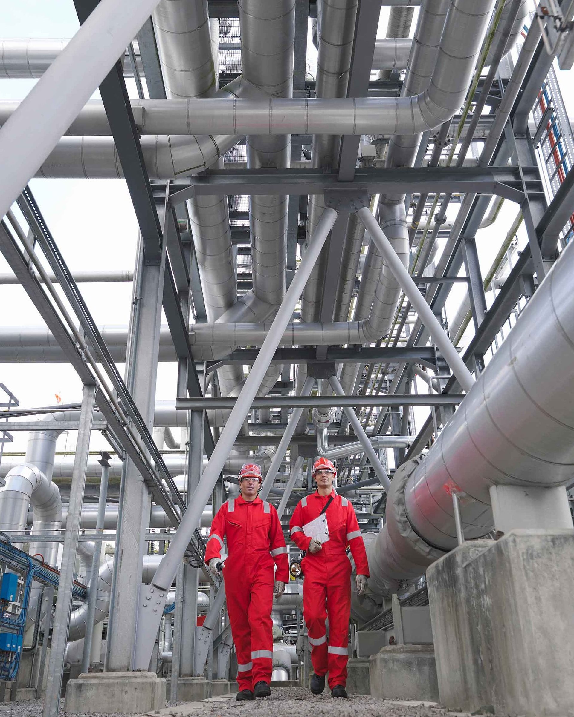 Zwei Personen mit Schutzhelmen und roten Jacken in einer Industrieanlage mit zahlreichen Rohren und Metallstrukturen