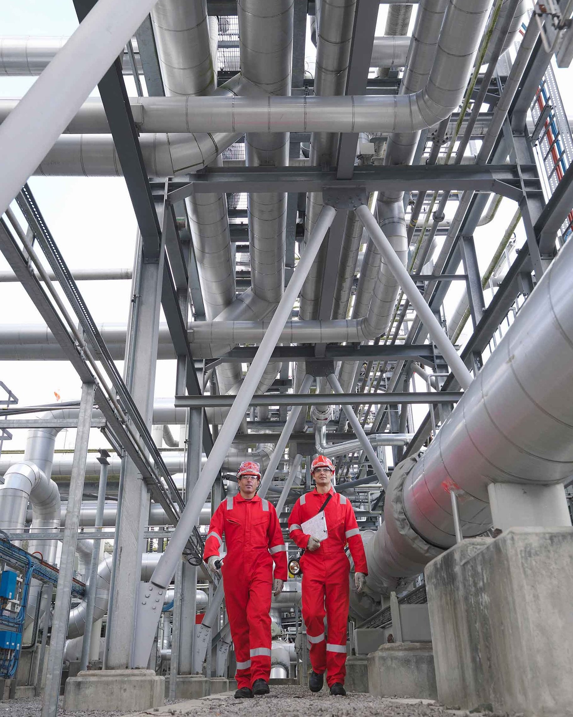 Two persons wearing hard hats and red jackets in an industrial plant with numerous pipes and metal structures