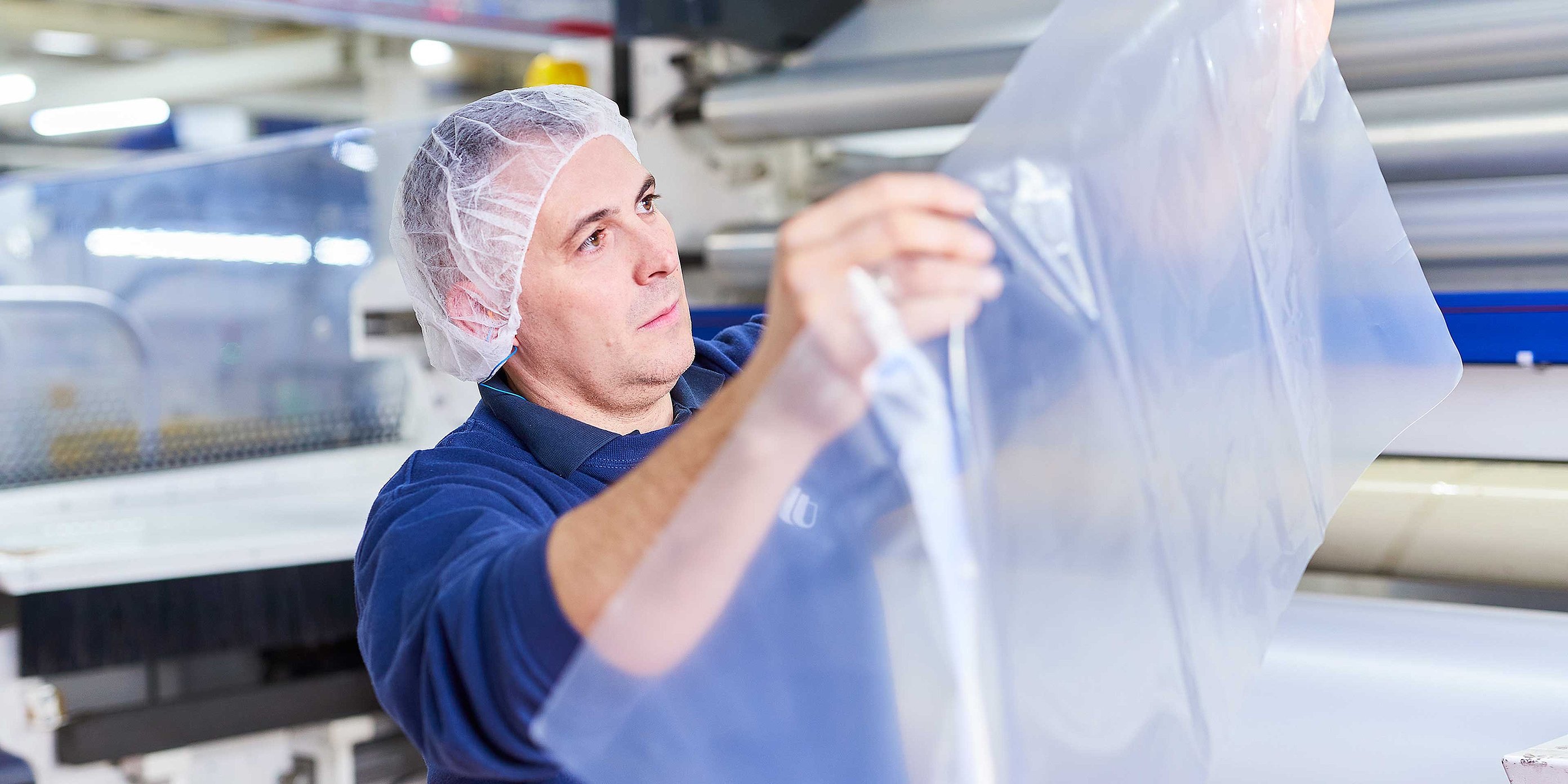 An employee wearing dark blue work clothing and a hair net is holding a plastic film in his hands