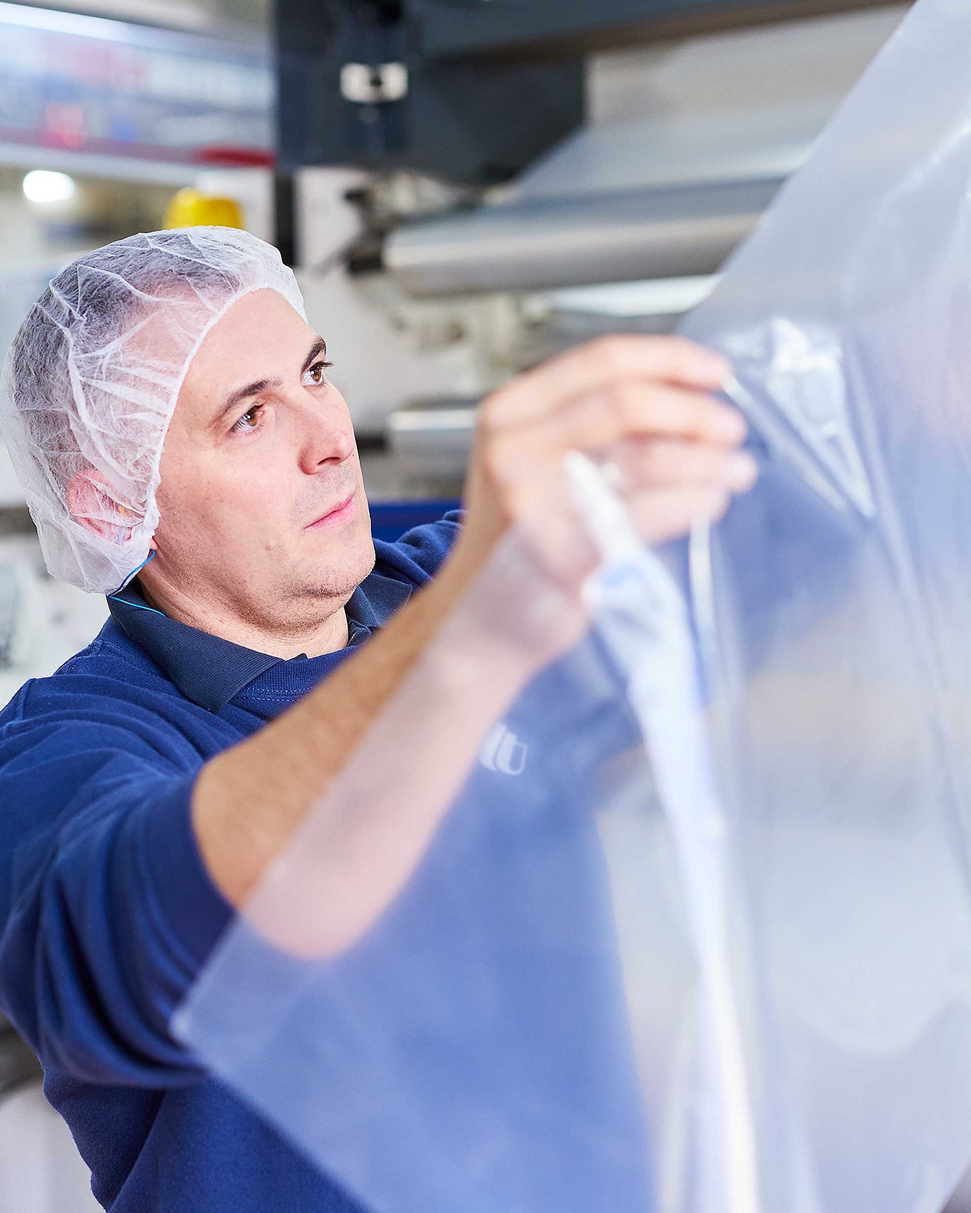 An employee wearing dark blue work clothing and a hair net is holding a plastic film in his hands