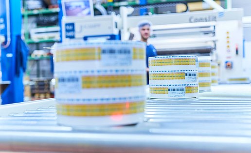 Produced plastic packaging on a reel is running along a conveyor belt. An employee can be seen in the background