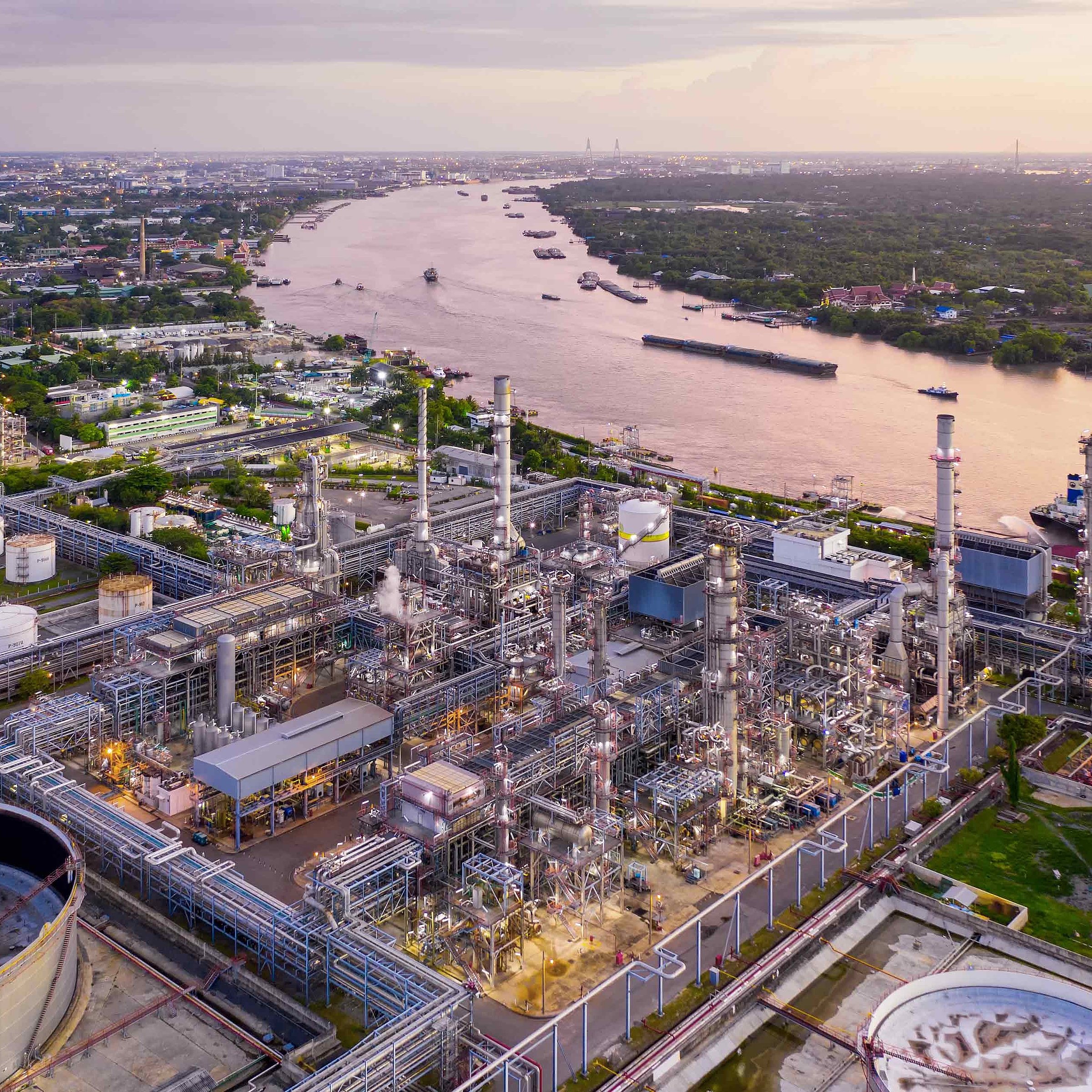 Chemical plant with numerous pipes and towers by a river with several ships at sunset