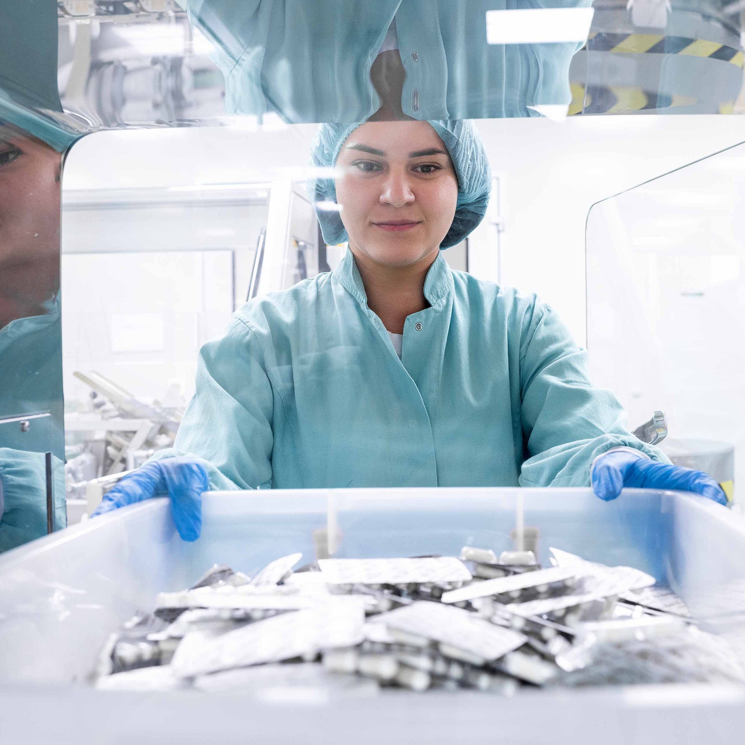 Eine Frau mit Schutzkleidung und Haube hält eine Box mit Tablettenblistern in einer Produktionsanlage