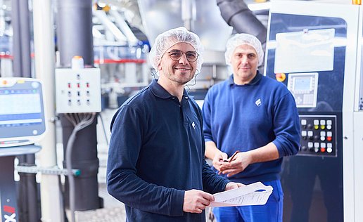Two employees in dark blue work clothing are standing in front of a machine in the production department. They are both wearing hair nets; one employee is holding a piece of paper in his hand