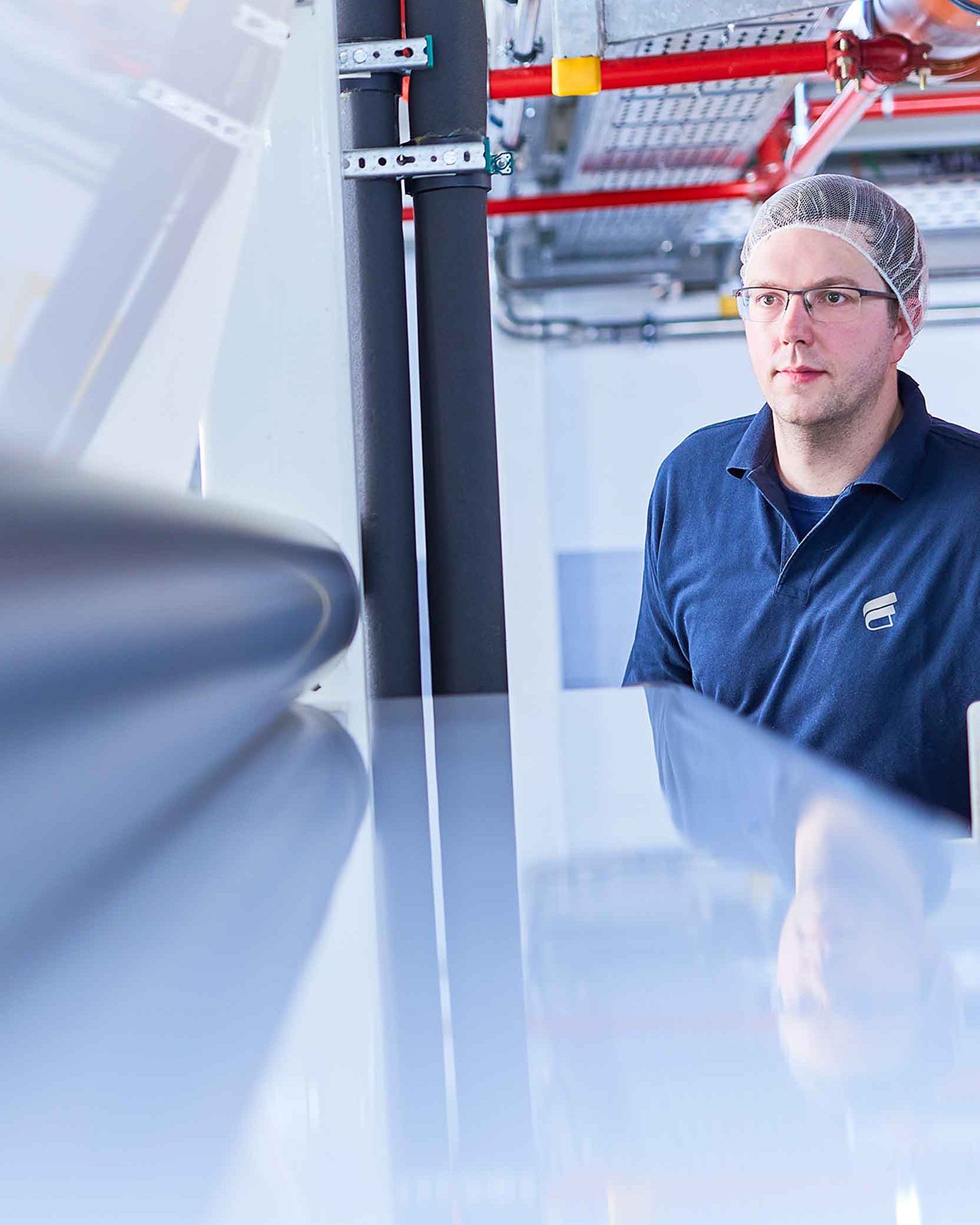 Ein Mitarbeiter mit Bischof+Klein Arbeitskleidung trägt Schutzhaube und Brille und schaut in eine Maschine in der Produktion 
