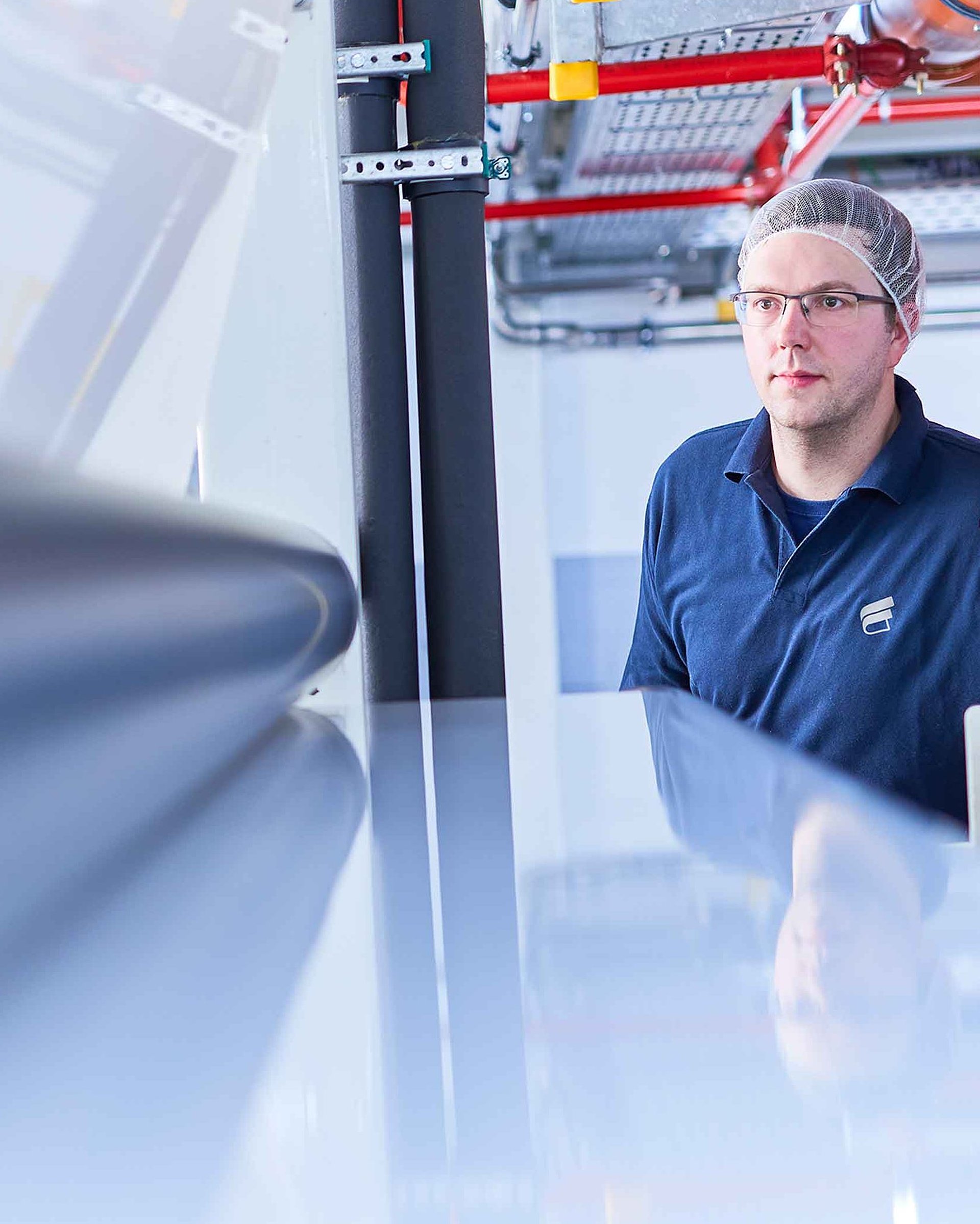 Un collaborateur en vêtement de travail de Bischof+Klein porte une coiffe et des lunettes de protection et regarde dans une machine de la production 