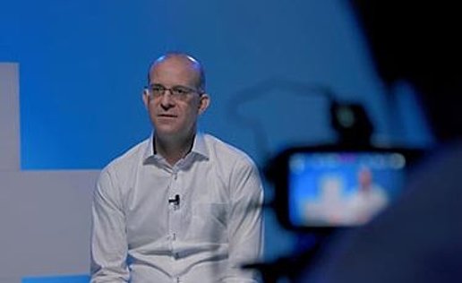Our CSO Erik Edelmann is wearing a white shirt and sitting behind a camera. He is currently being interviewed and the interview is being filmed