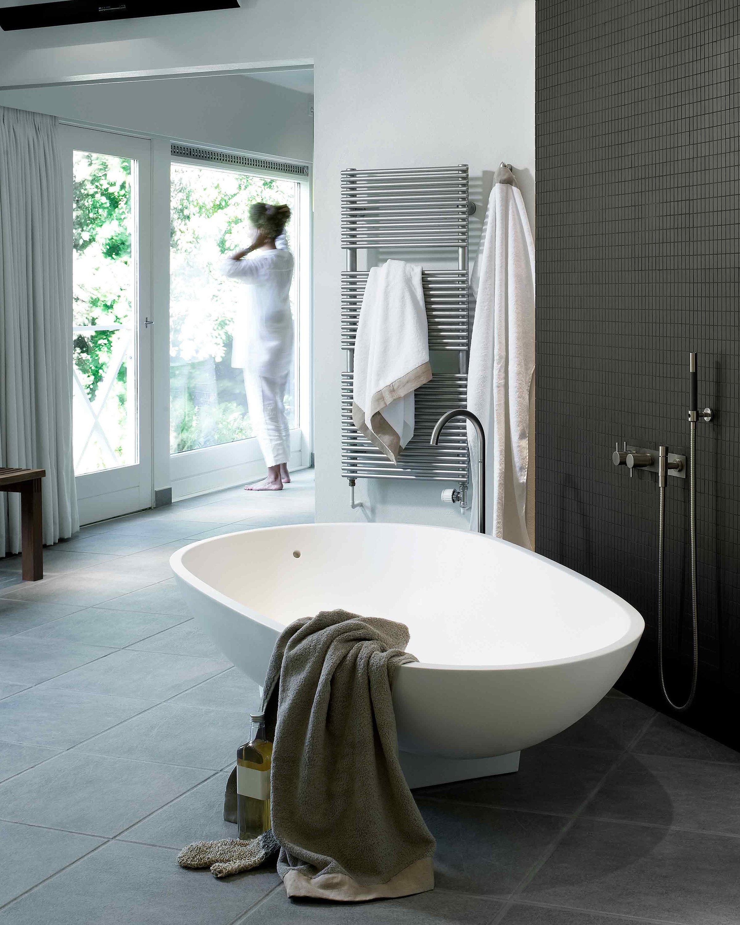 A bathroom can be seen. The focus is on a bathtub on which a brown towel is lying. A shower fitting and a towel rail on which two white towels are hanging are shown in the background.