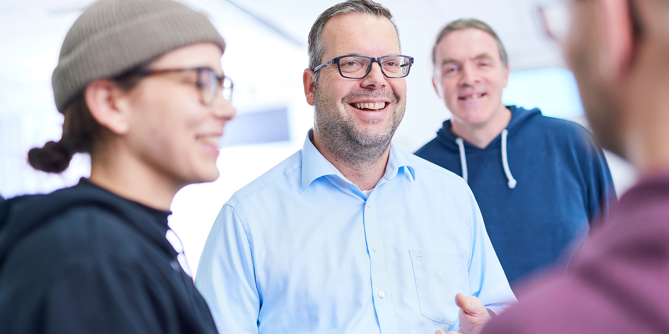 Four employees are standing together and talking. One of them is looking directly into the camera. He is wearing black glasses.