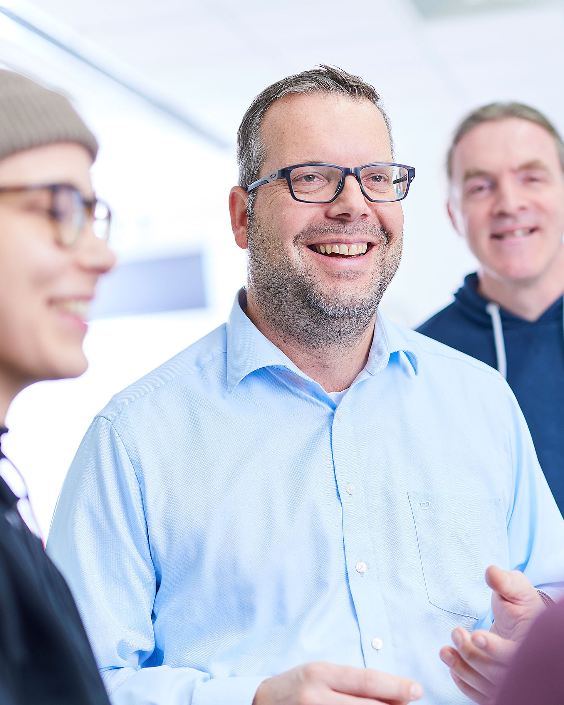 Four employees are standing together and talking. One of them is looking directly into the camera. He is wearing black glasses.