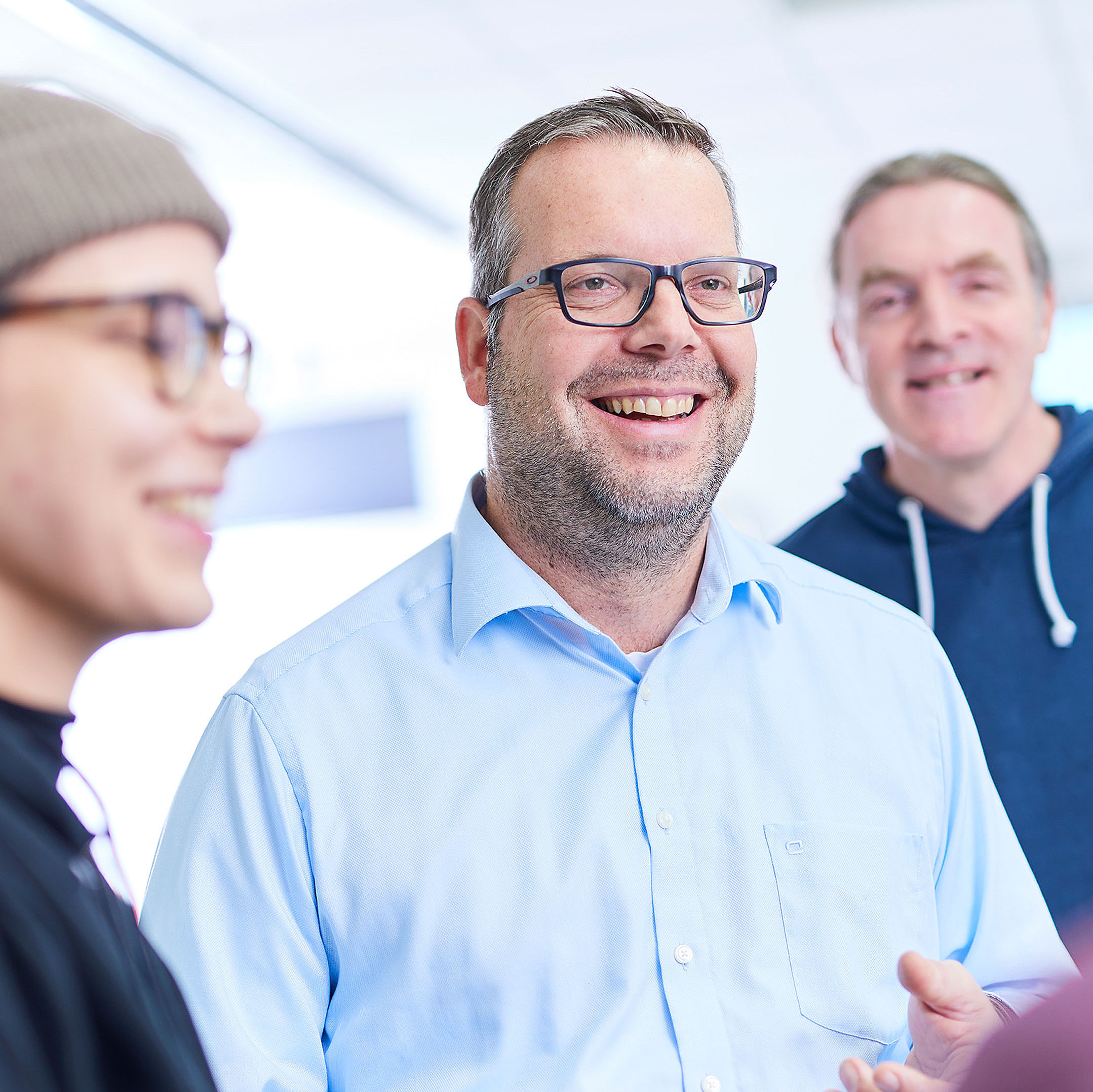 Four employees are standing together and talking. One of them is looking directly into the camera. He is wearing black glasses.