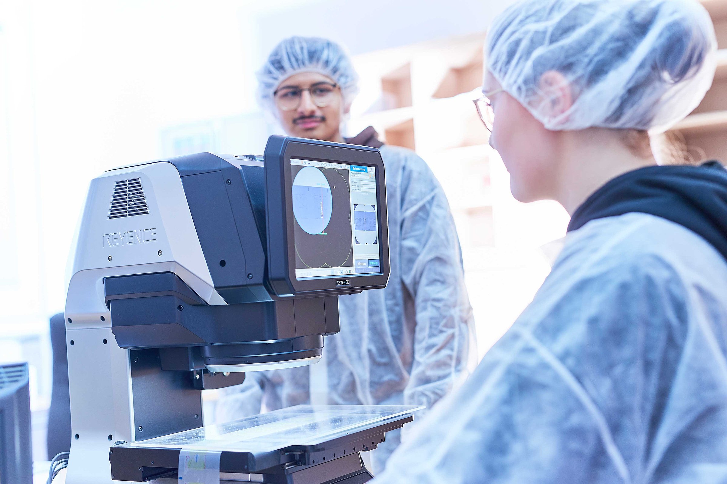 Two people in protective gear are operating a measuring device with a screen in a laboratory