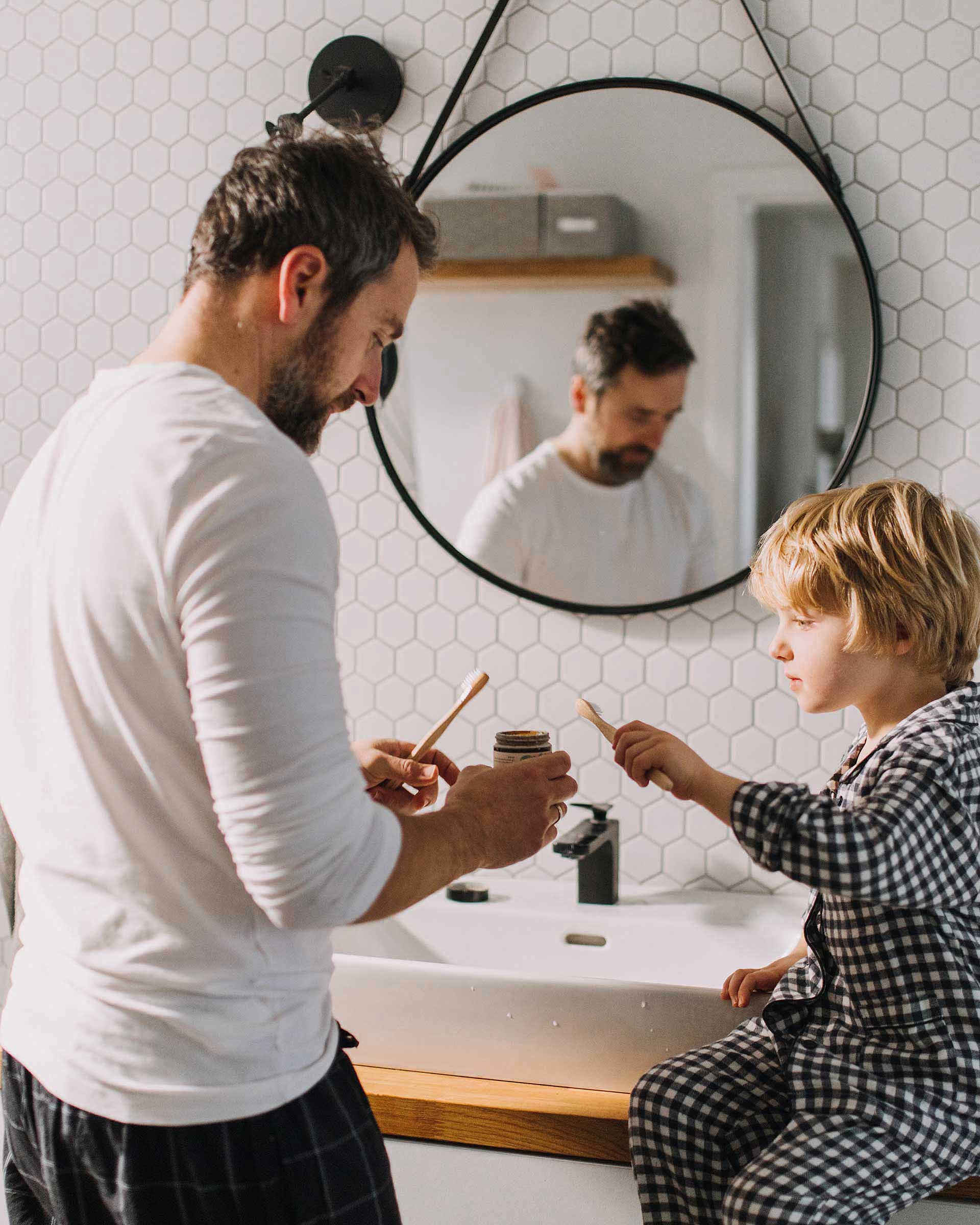 A father is standing at the sink in the bathroom with his son. Both are holding a toothbrush in their hands. The father is holding a tube of toothpaste in his hand. White tiles and a mirror can be seen in the background