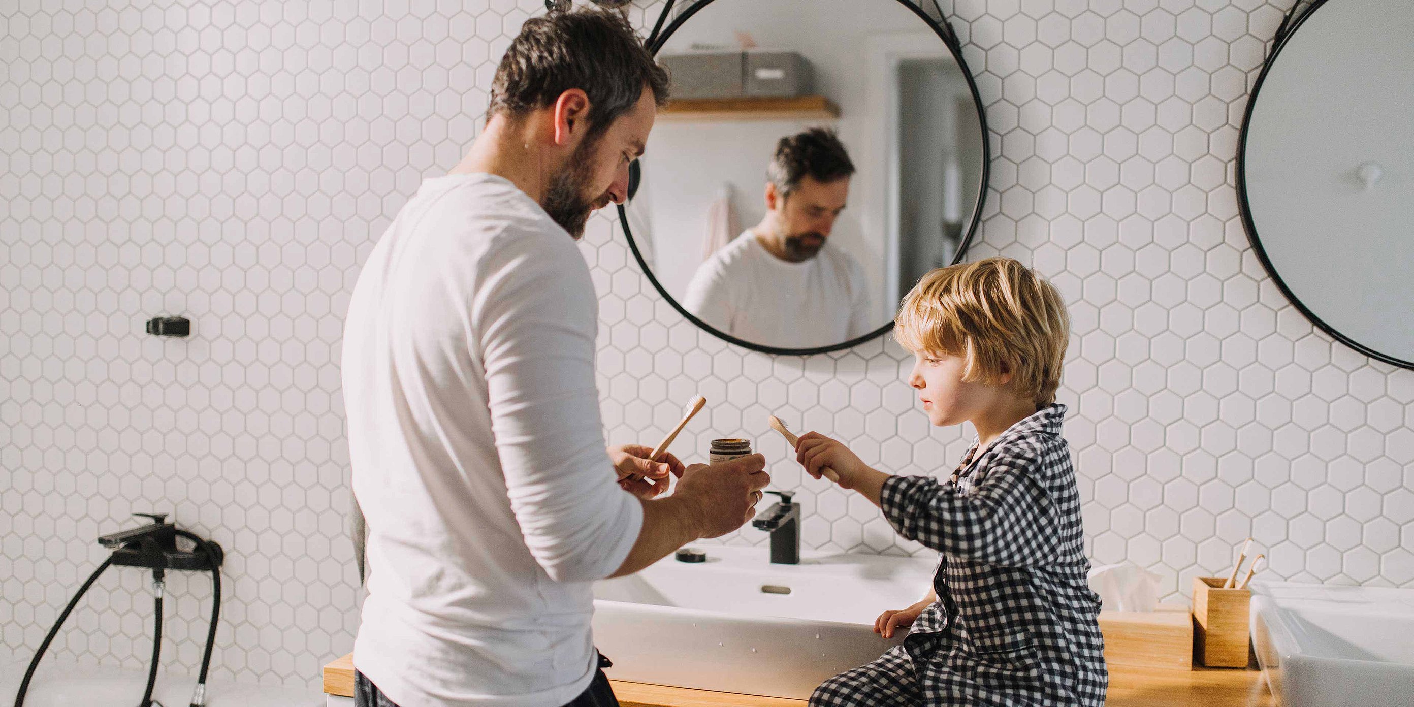 A father is standing at the sink in the bathroom with his son. Both are holding a toothbrush in their hands. The father is holding a tube of toothpaste in his hand. White tiles and a mirror can be seen in the background