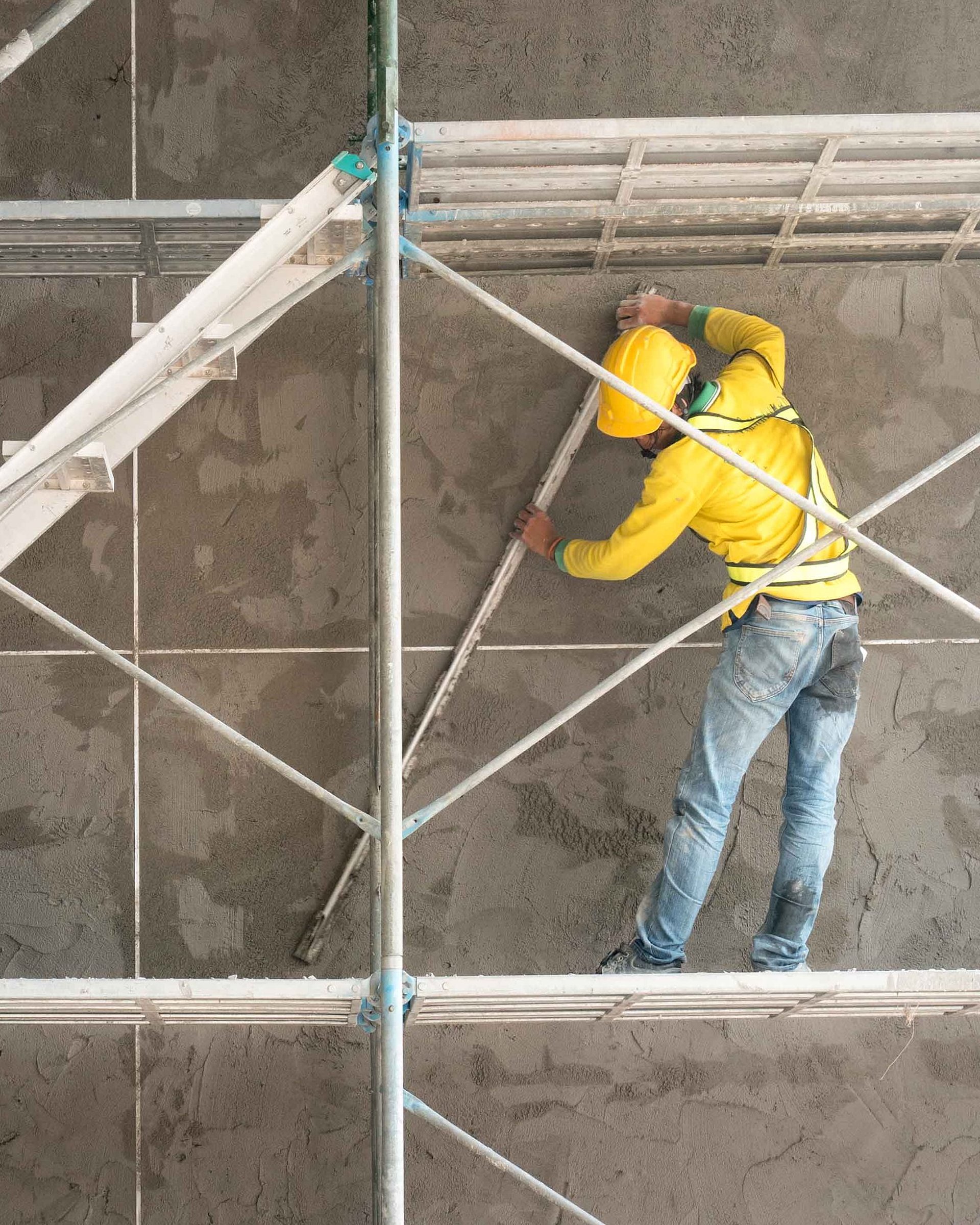 A construction worker wearing yellow protective clothing and a helmet is working on scaffolding in front of a concrete wall