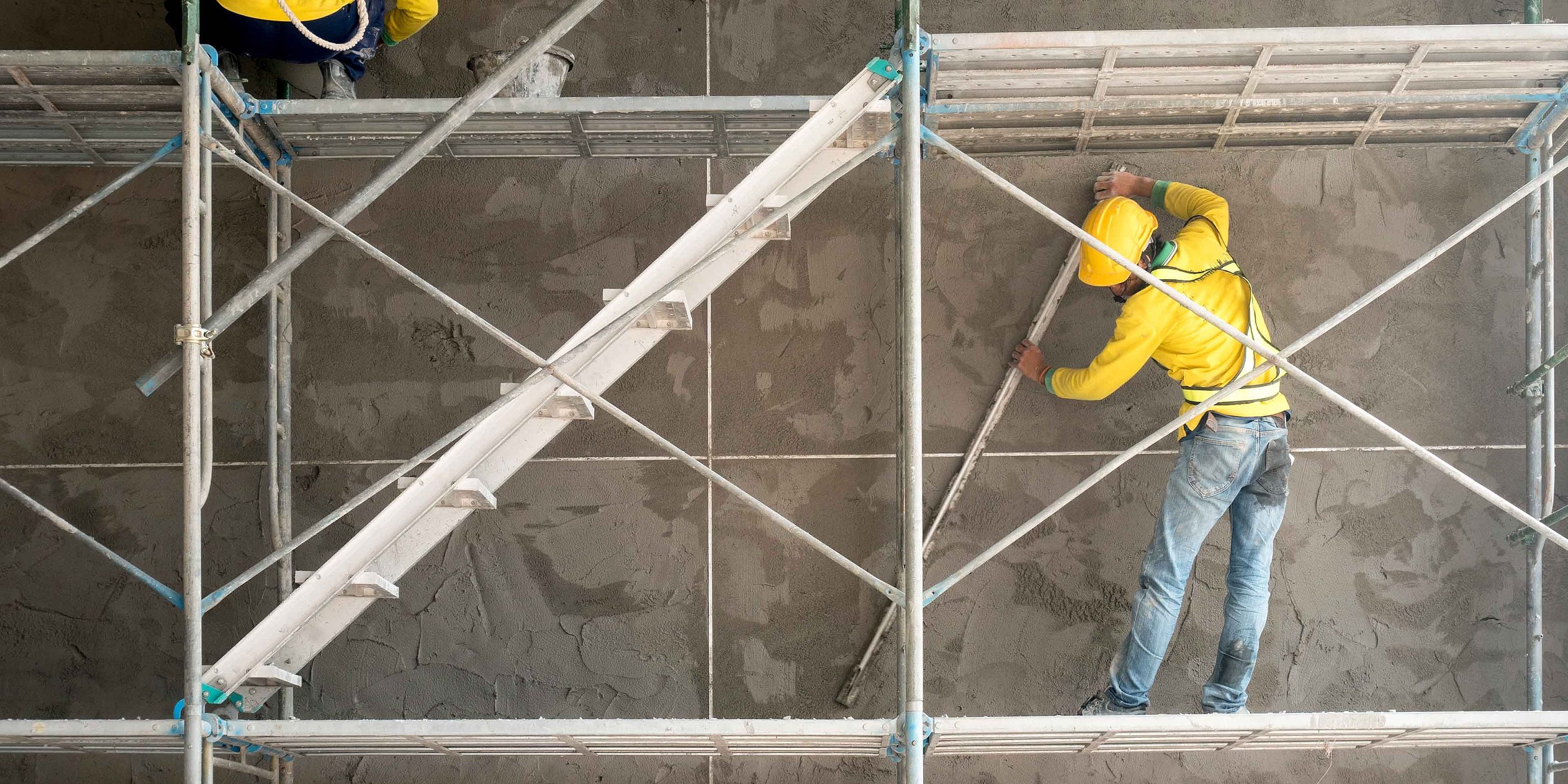 A construction worker wearing yellow protective clothing and a helmet is working on scaffolding in front of a concrete wall