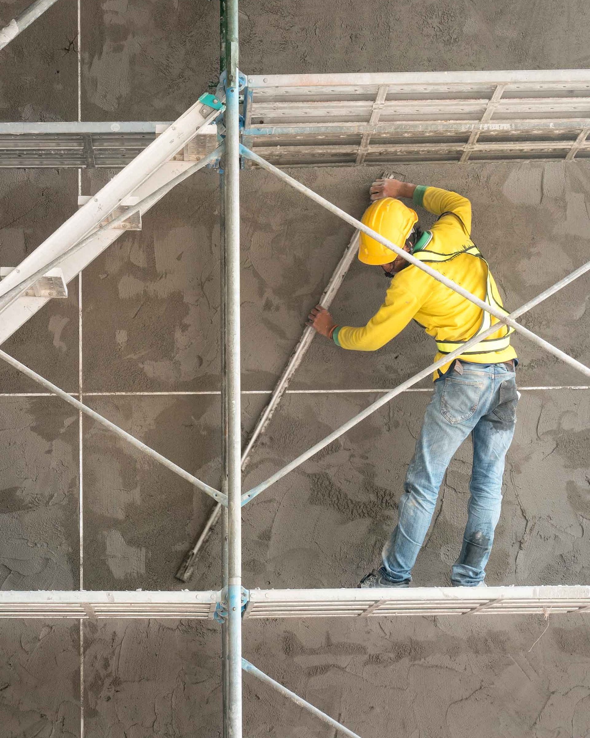 Pracownik budowlany w żółtej odzieży ochronnej i kasku pracujący na rusztowaniu przed betonową ścianą