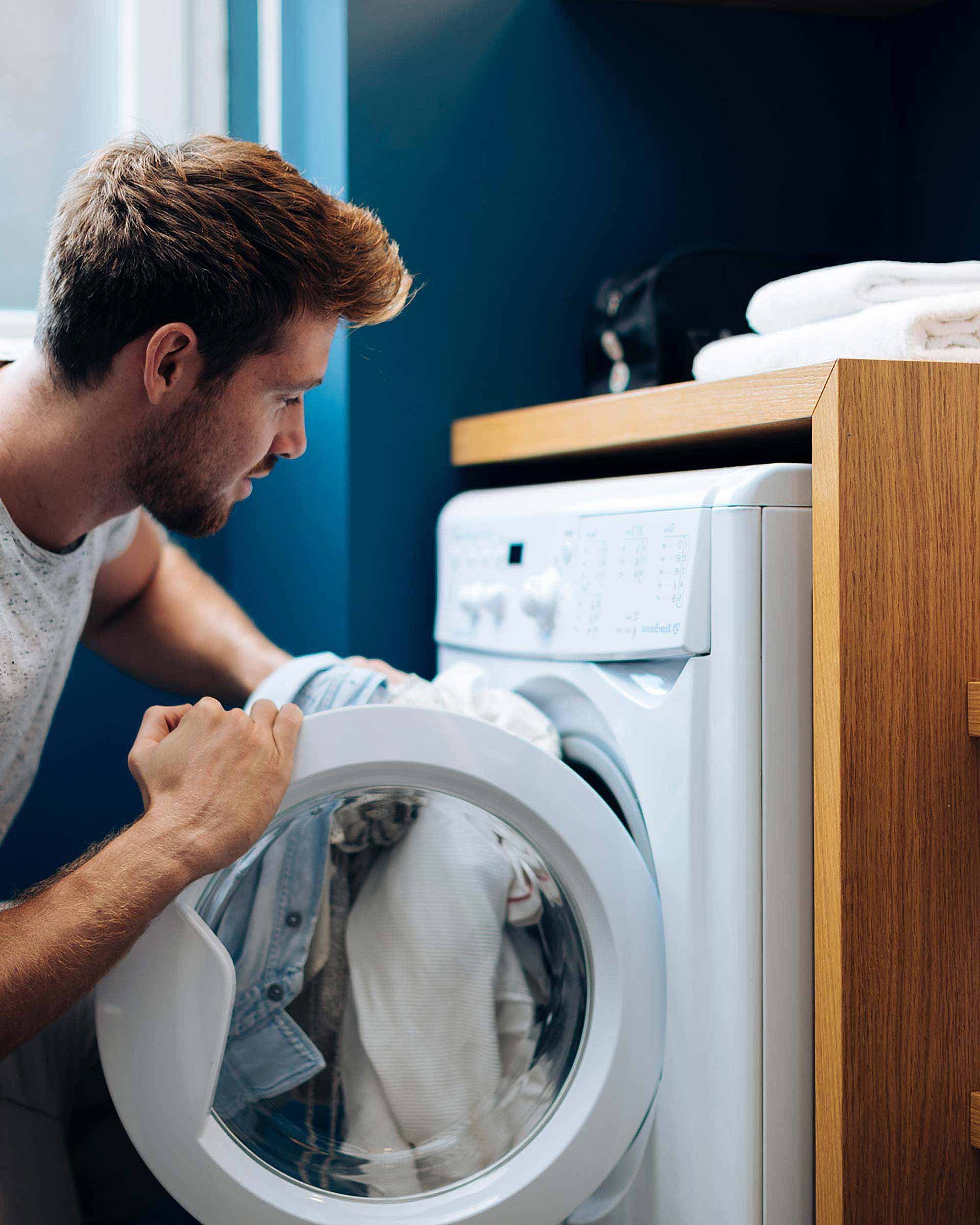 A man is loading his dirty laundry into a washing machine. A washbasin can be seen next to him
