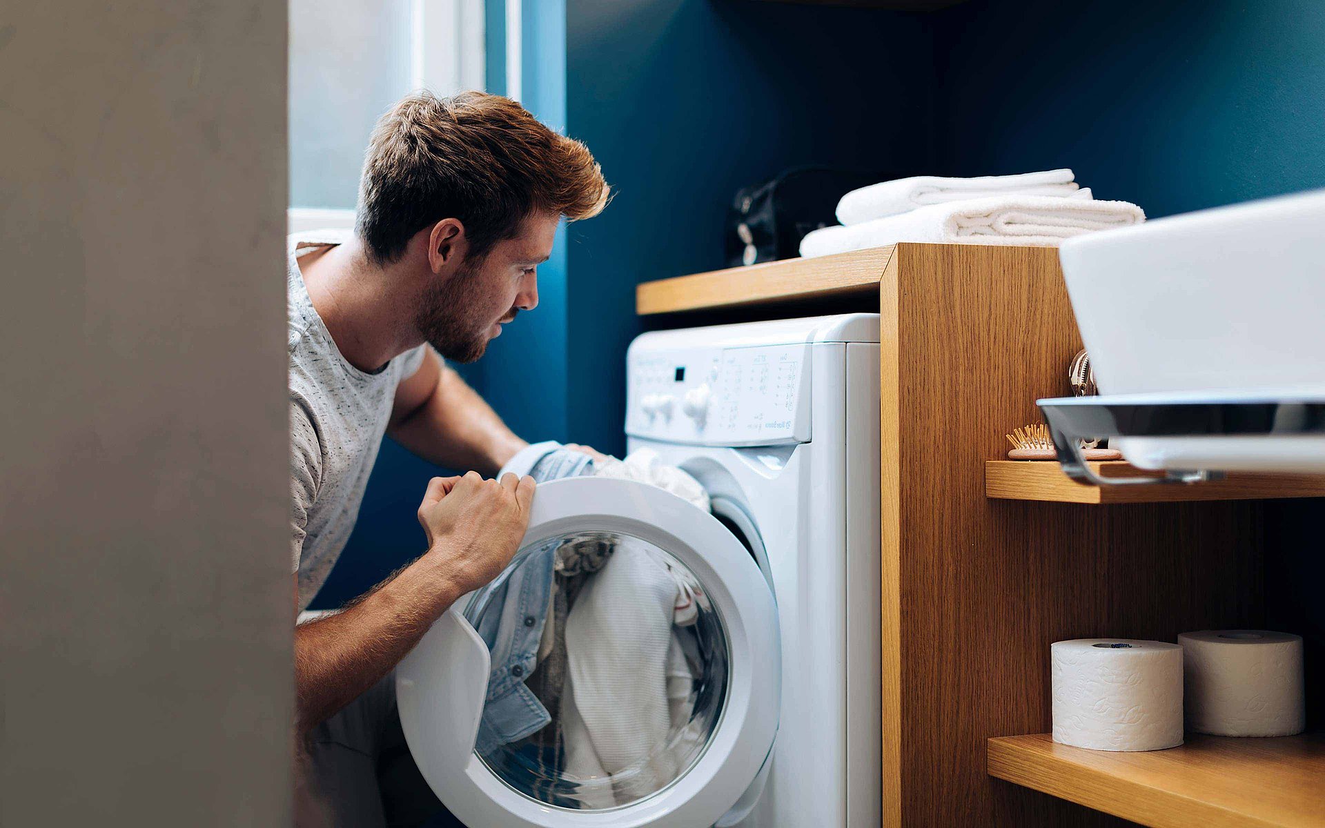 Un homme met son linge sale dans une machine à laver. A côté de lui, on peut voir un lavabo