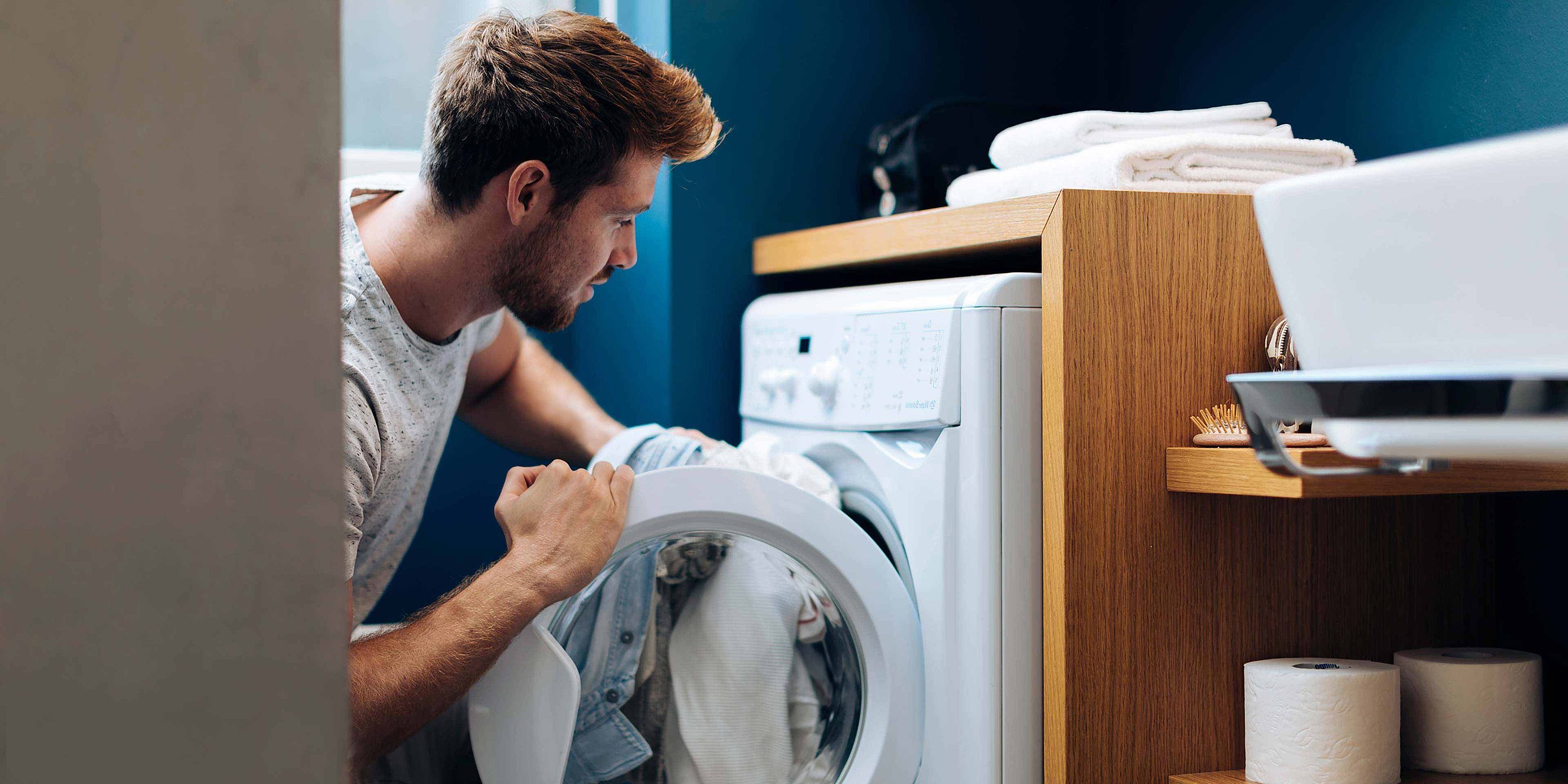 A man is loading his dirty laundry into a washing machine. A washbasin can be seen next to him
