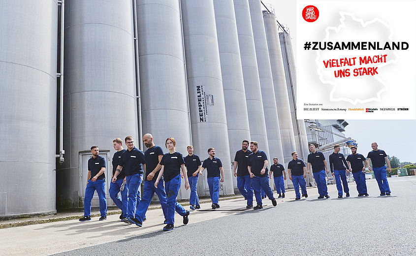 Kilku stażystów w ubraniach roboczych spaceruje wzdłuż silosów w hali produkcyjnej w Lengerich