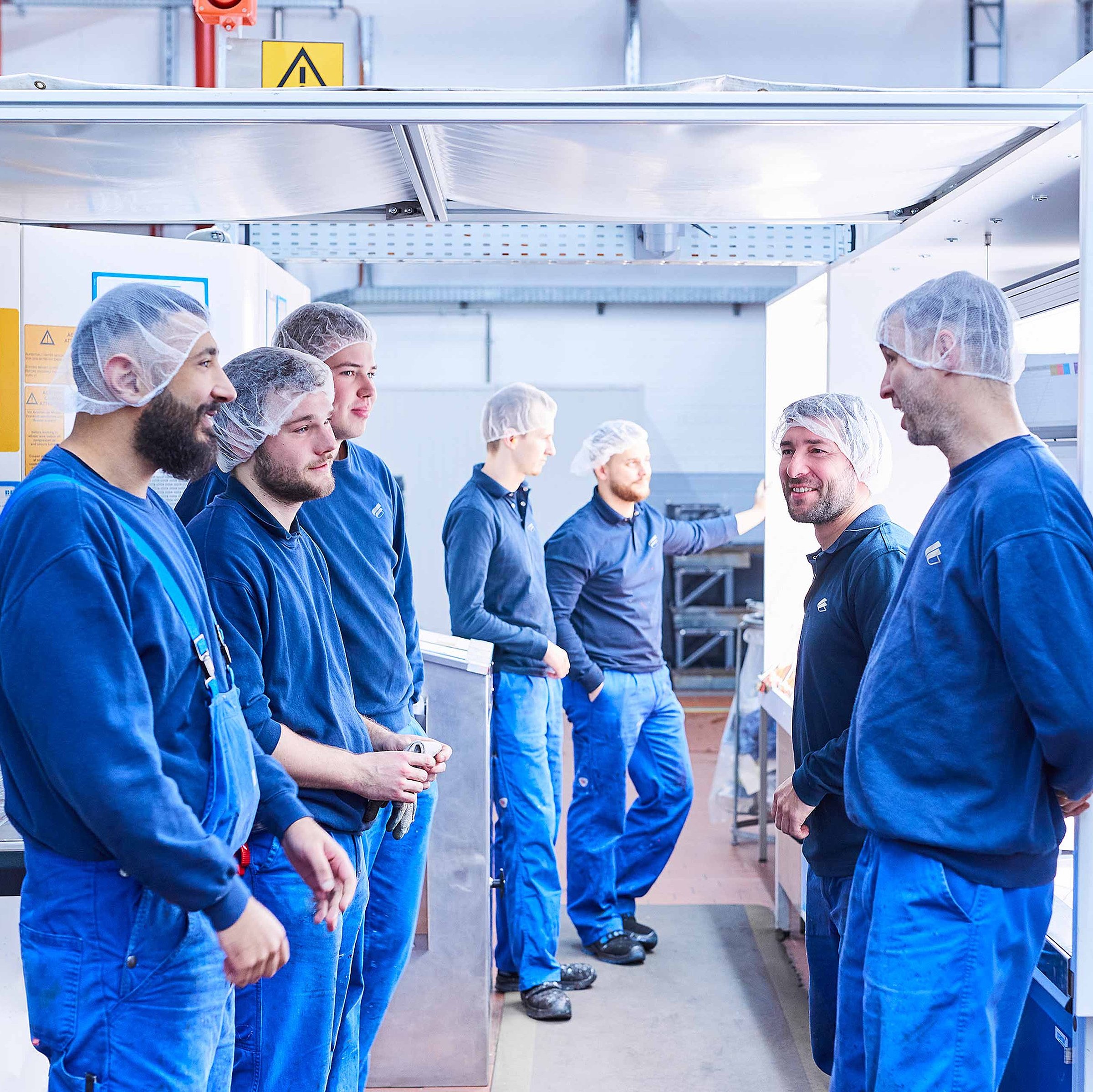 The picture shows seven Bischof+Klein employees. They are all wearing dark blue work clothing and hair nets and are standing in the production hall