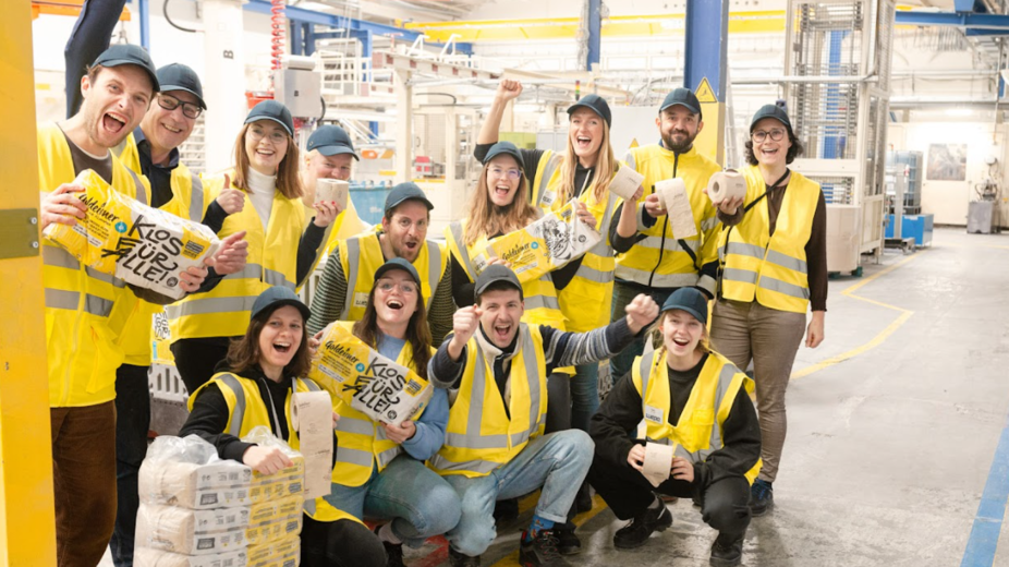  Bischof+Klein Mitarbeiterin zusammen mit Mitarbeitenden von Wildplastic, Goldeimer und Wepa in der Produktionshalle. Sie halten alle einen Toilettenpapierpackung von Goldeimer in der Hand