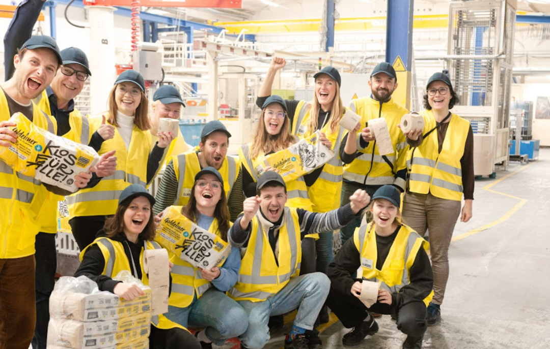  Bischof+Klein Mitarbeiterin zusammen mit Mitarbeitenden von Wildplastic, Goldeimer und Wepa in der Produktionshalle. Sie halten alle einen Toilettenpapierpackung von Goldeimer in der Hand