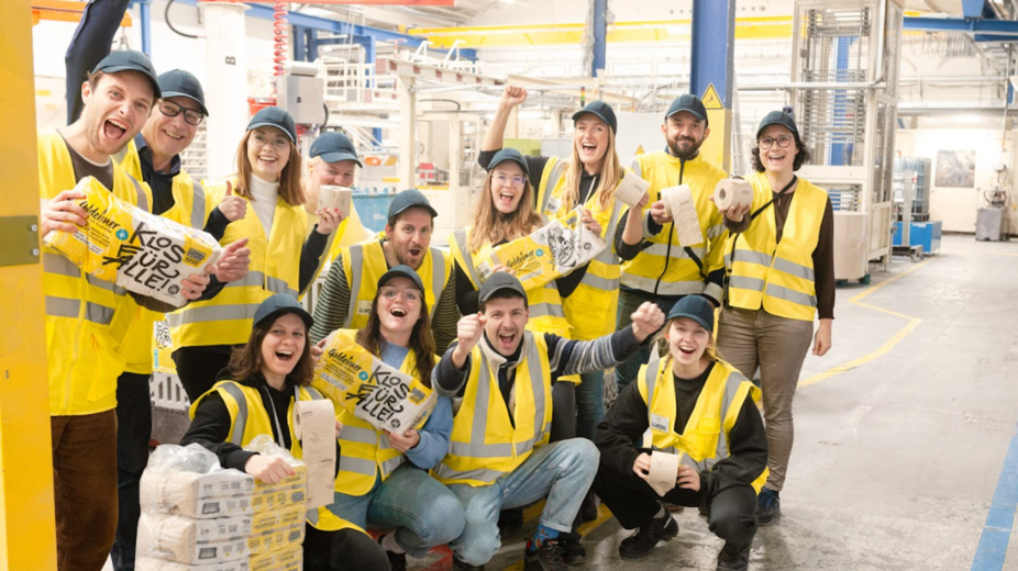 Des collaborateurs de Bischof+Klein avec des collaborateurs de Wildplastic, Goldeimer et Wepa dans le hall de production. Ils tiennent tous un paquet de papier toilette de Goldeimer dans la main