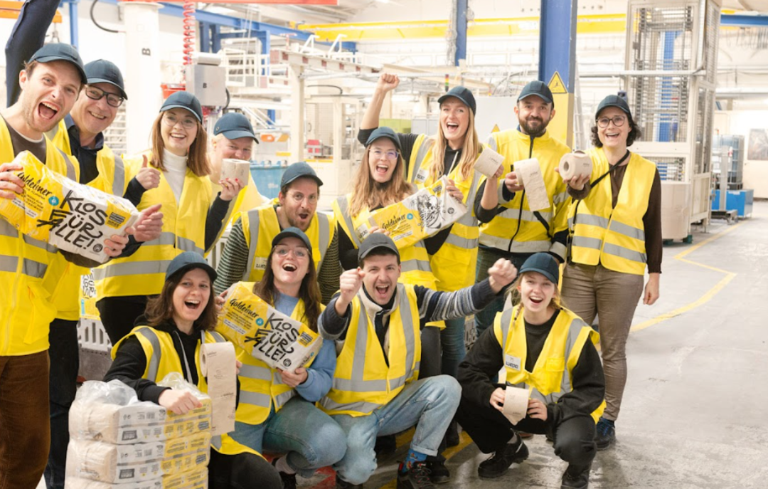 Des collaborateurs de Bischof+Klein avec des collaborateurs de Wildplastic, Goldeimer et Wepa dans le hall de production. Ils tiennent tous un paquet de papier toilette de Goldeimer dans la main