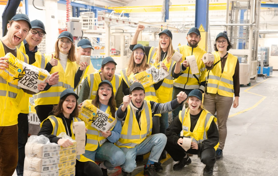  Bischof+Klein Mitarbeiterin zusammen mit Mitarbeitenden von Wildplastic, Goldeimer und Wepa in der Produktionshalle. Sie halten alle einen Toilettenpapierpackung von Goldeimer in der Hand