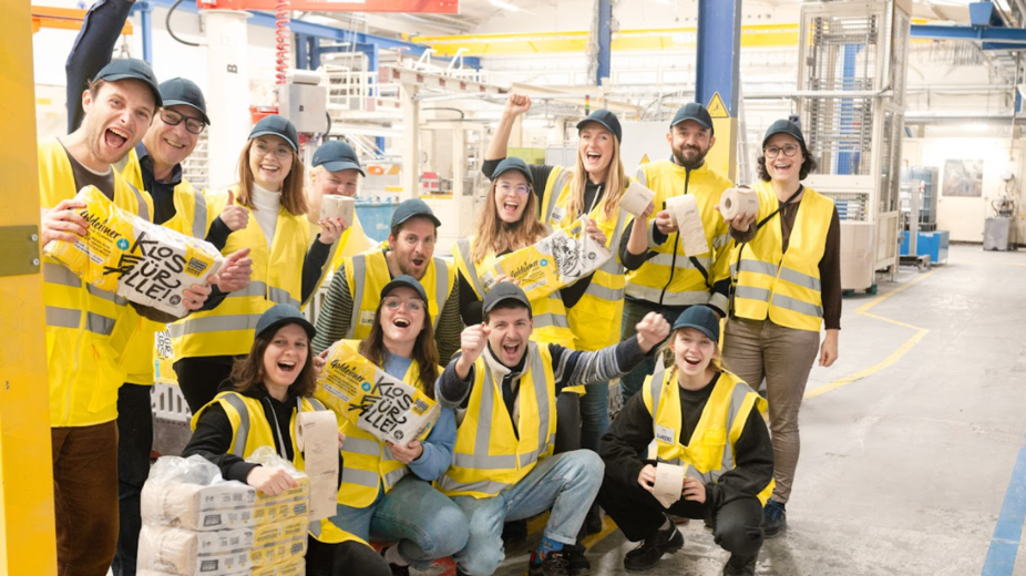  Bischof+Klein Mitarbeiterin zusammen mit Mitarbeitenden von Wildplastic, Goldeimer und Wepa in der Produktionshalle. Sie halten alle einen Toilettenpapierpackung von Goldeimer in der Hand