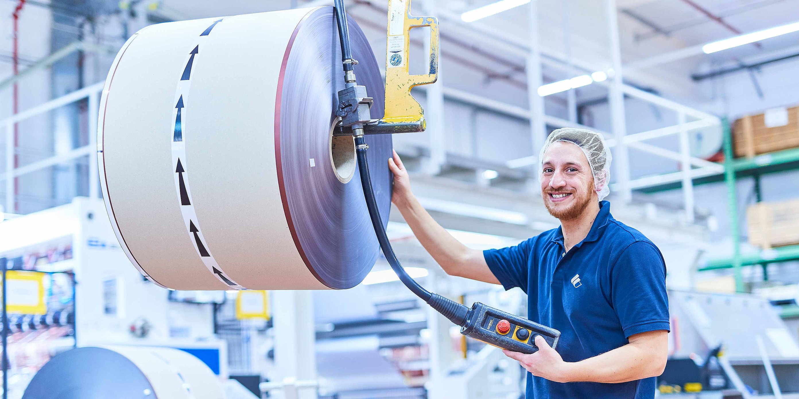 Ein Mitarbeiter in Bischof+Klein Arbeitskleidung mit Haarnetz steht neben einer produzierten Rolle mit Kunststofffolie und hält ein Bedienpanel in der Hand. 