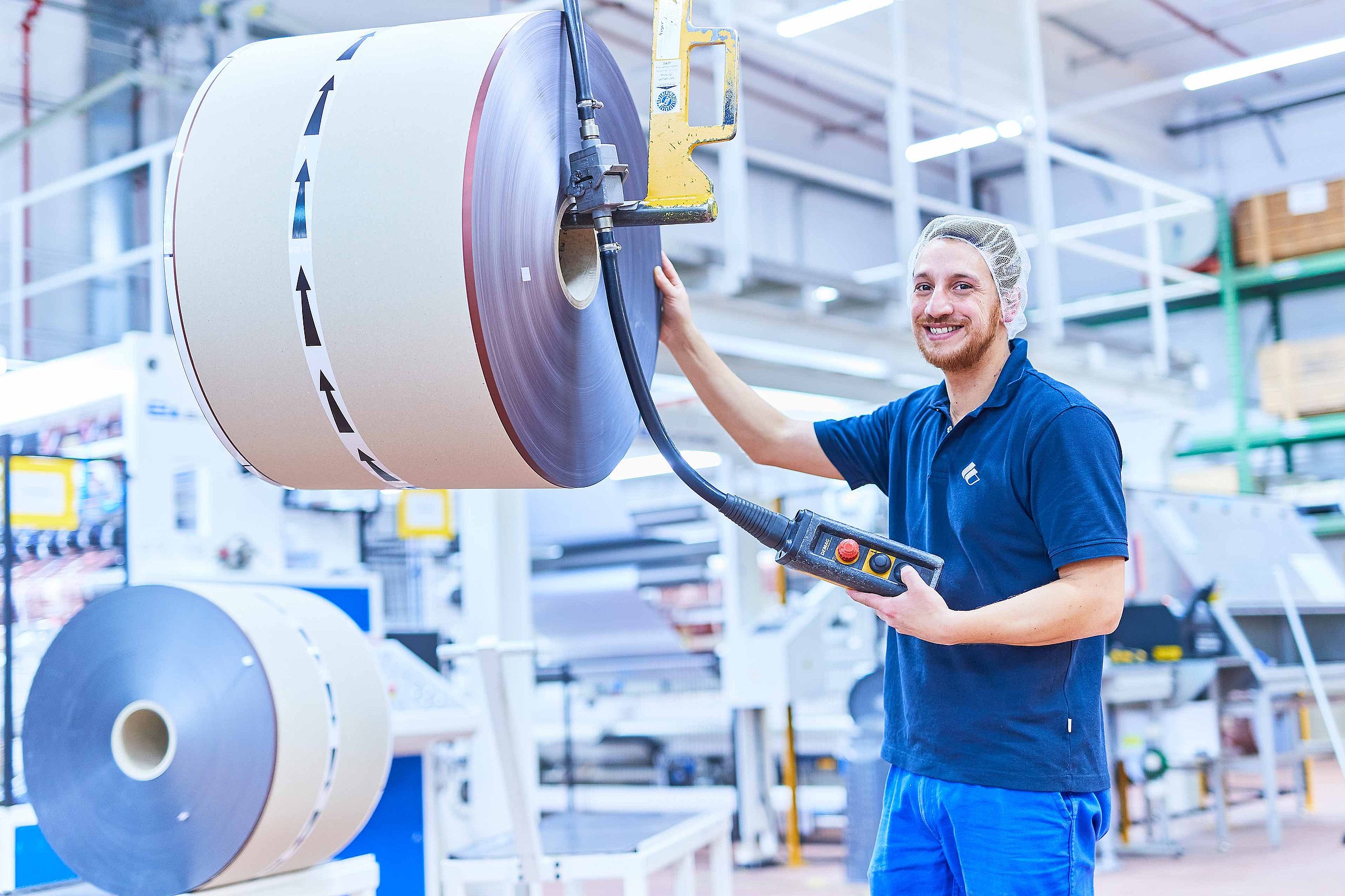 An employee wearing Bischof+Klein work clothing and a hair net is standing next to a produced reel of plastic film and holds a control panel in his hand. 