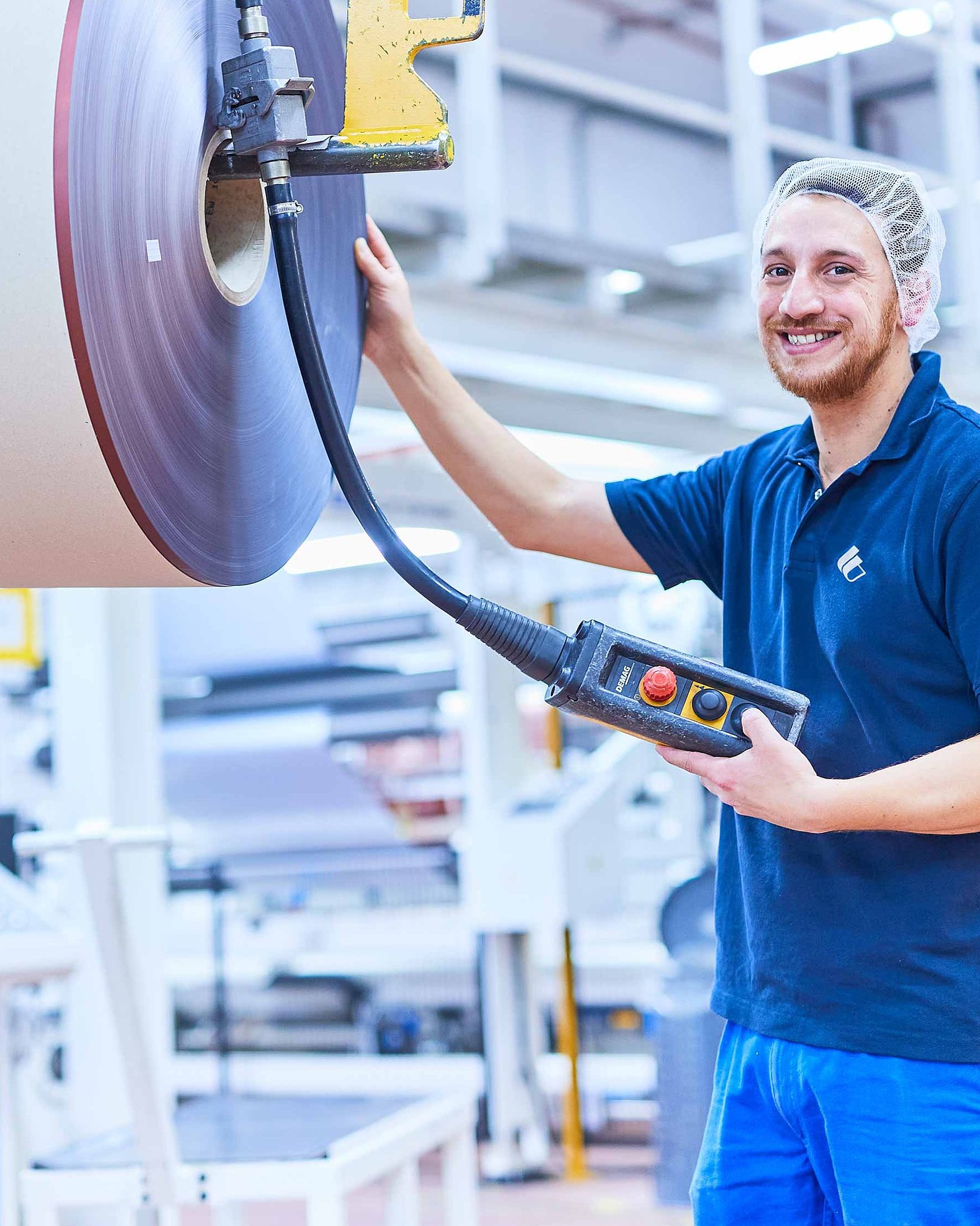 Ein Mitarbeiter in Bischof+Klein Arbeitskleidung mit Haarnetz steht neben einer produzierten Rolle mit Kunststofffolie und hält ein Bedienpanel in der Hand. 