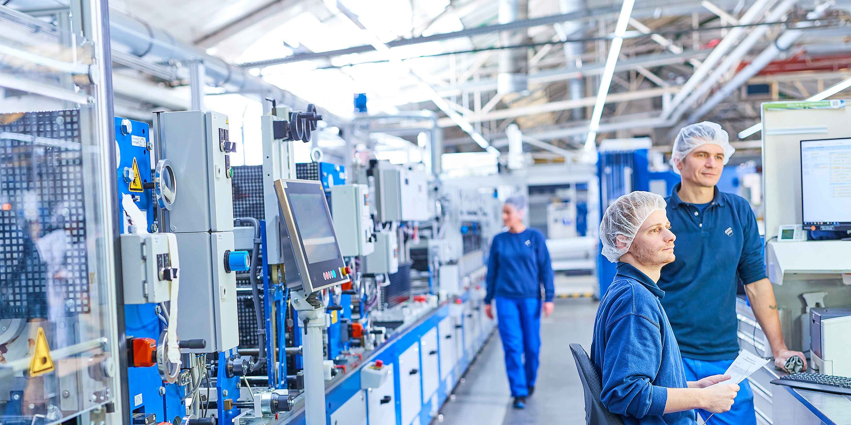 Two employees in Bischof+Klein work clothing with hair nets in the production department. They are standing or sitting next to a machine