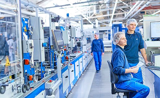 Two employees in Bischof+Klein work clothing with hair nets in the production department. They are standing or sitting next to a machine