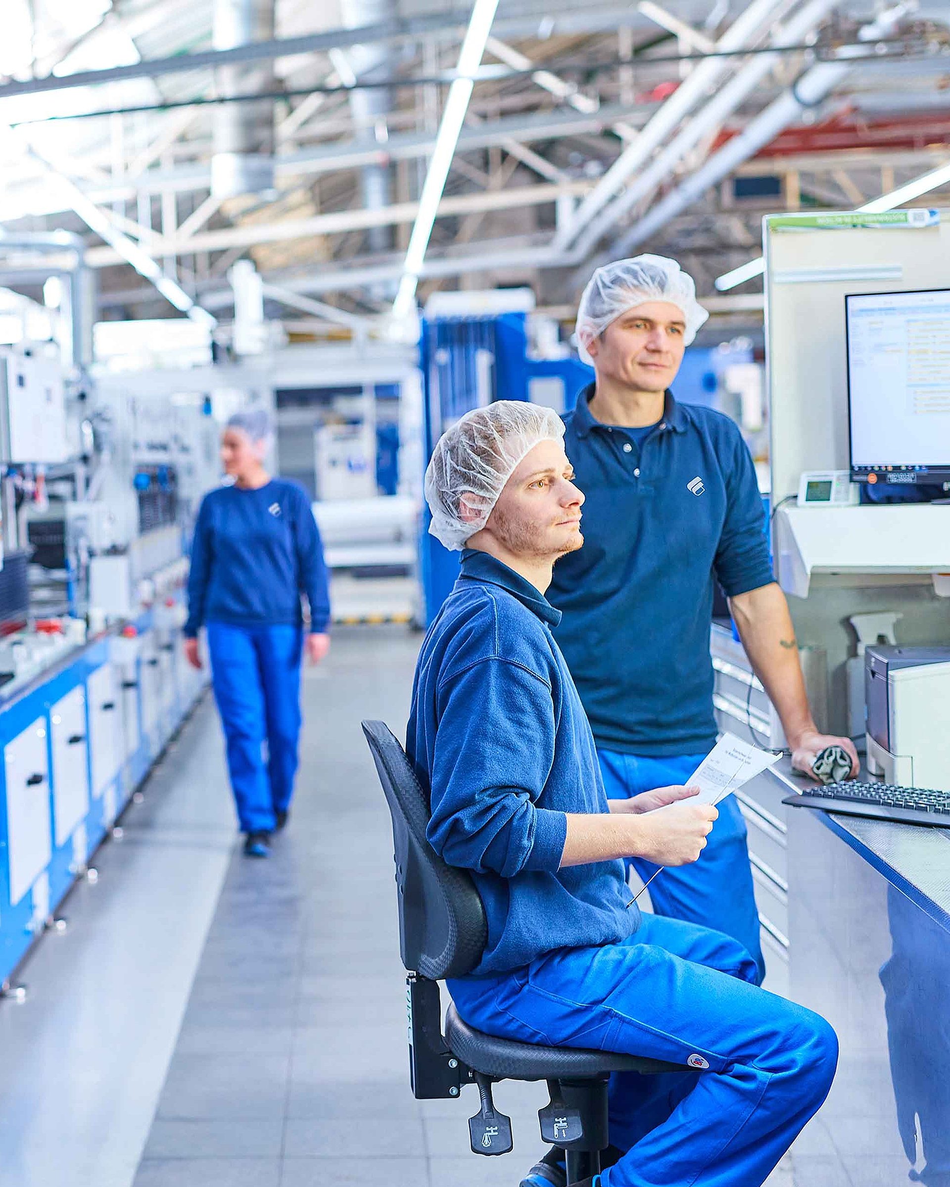 Deux collaborateurs en vêtements de travail de Bischof+Klein portant une résille à la production. Ils se tiennent debout ou assis à côté d’une machine